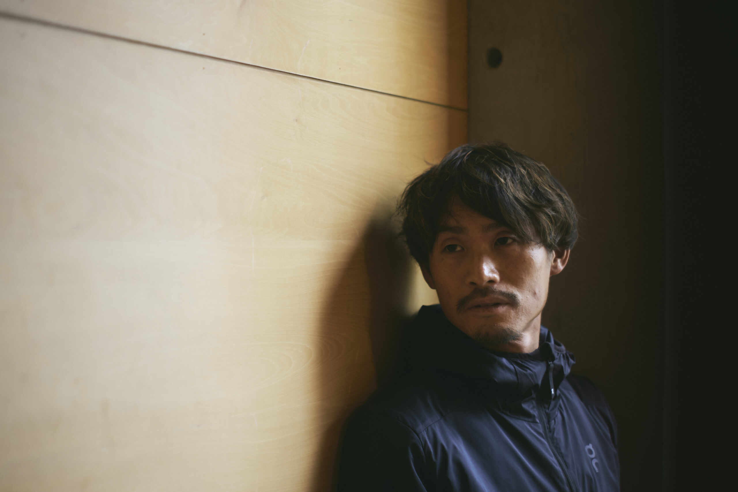 壁を背に遠くを見つめている横田さんの写真