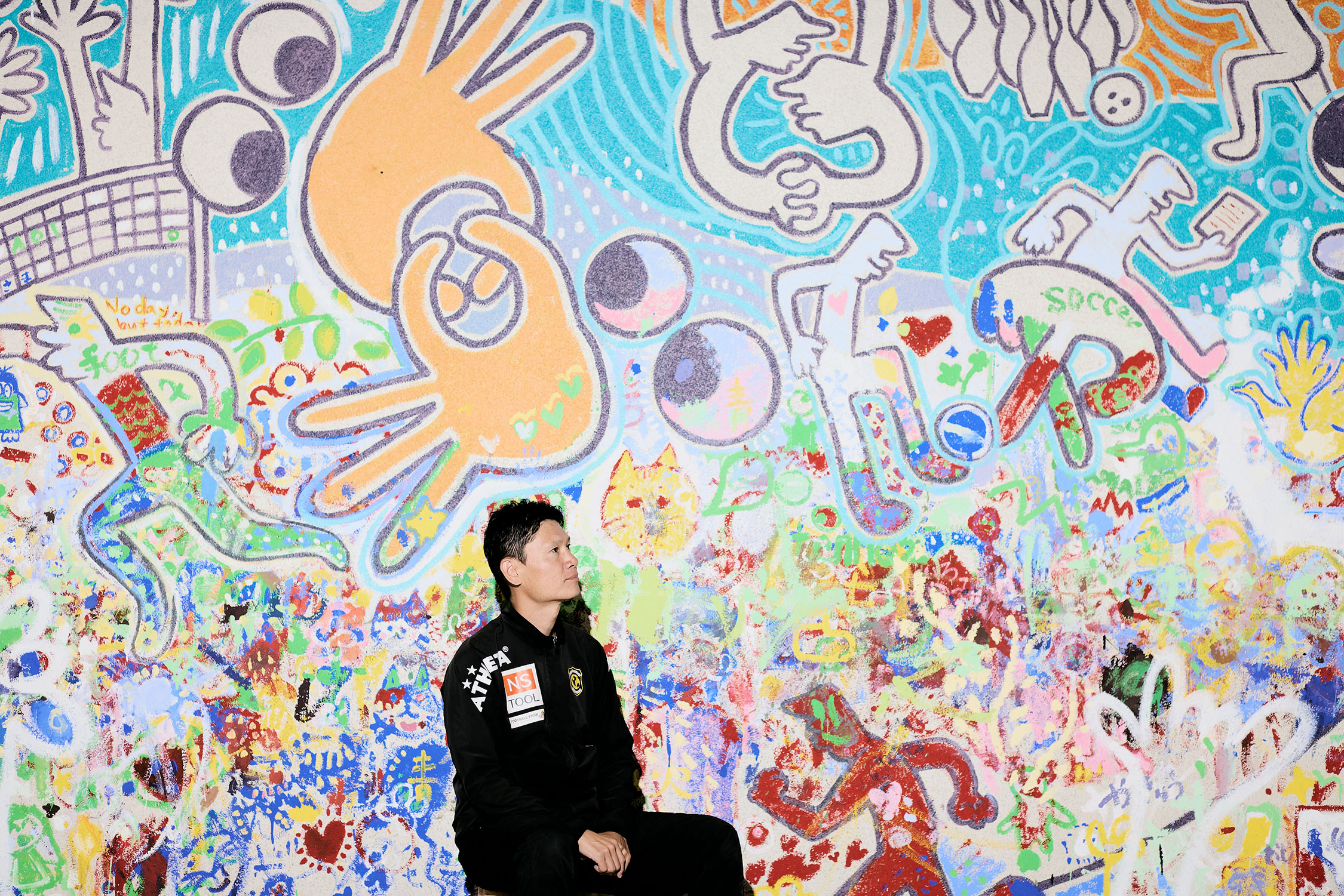 手話アートの前に座って斜め上を見つめている植松さんの写真
