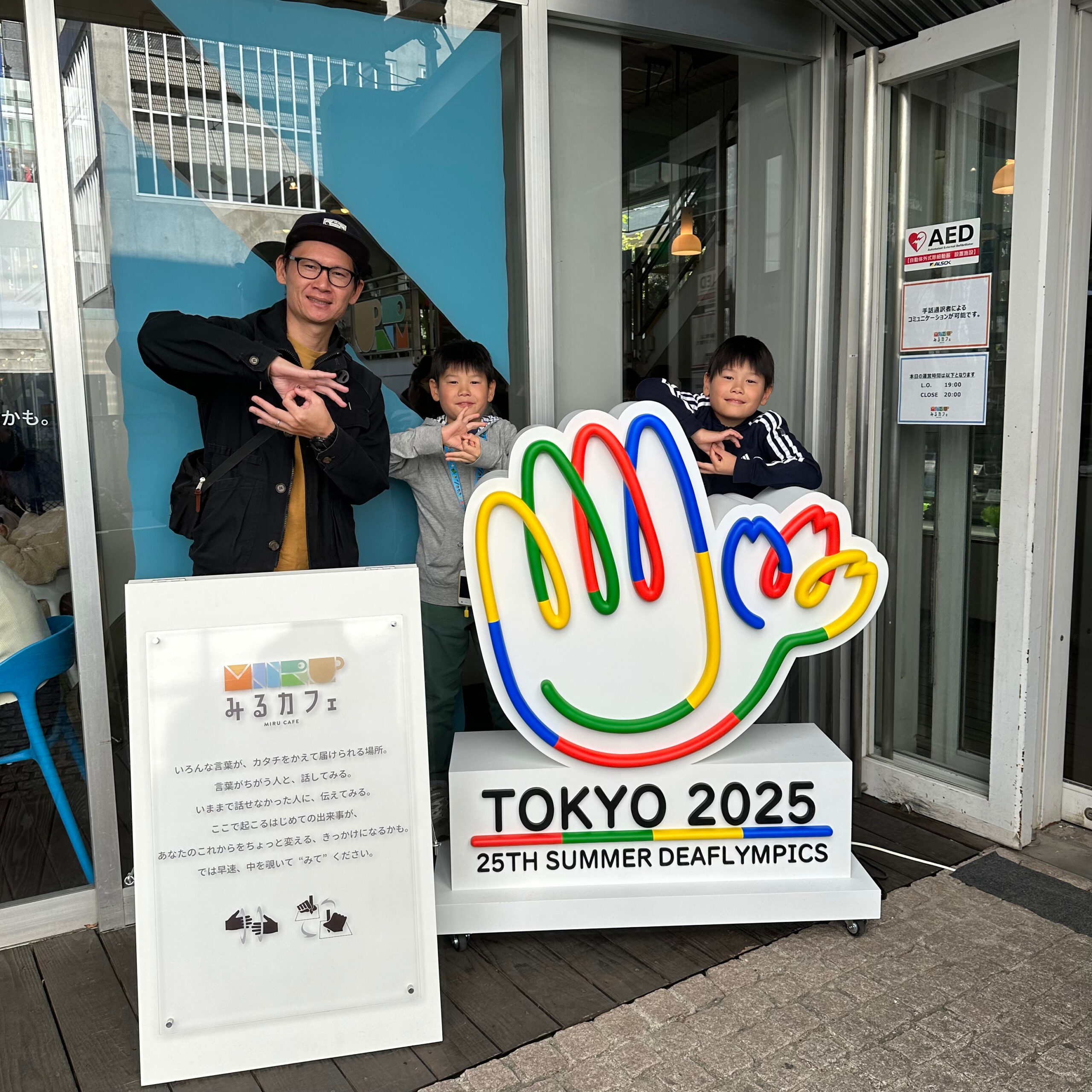 みるカフェ看板とデフリンピックのパネル前で子どもと一緒に写る植松さんの写真