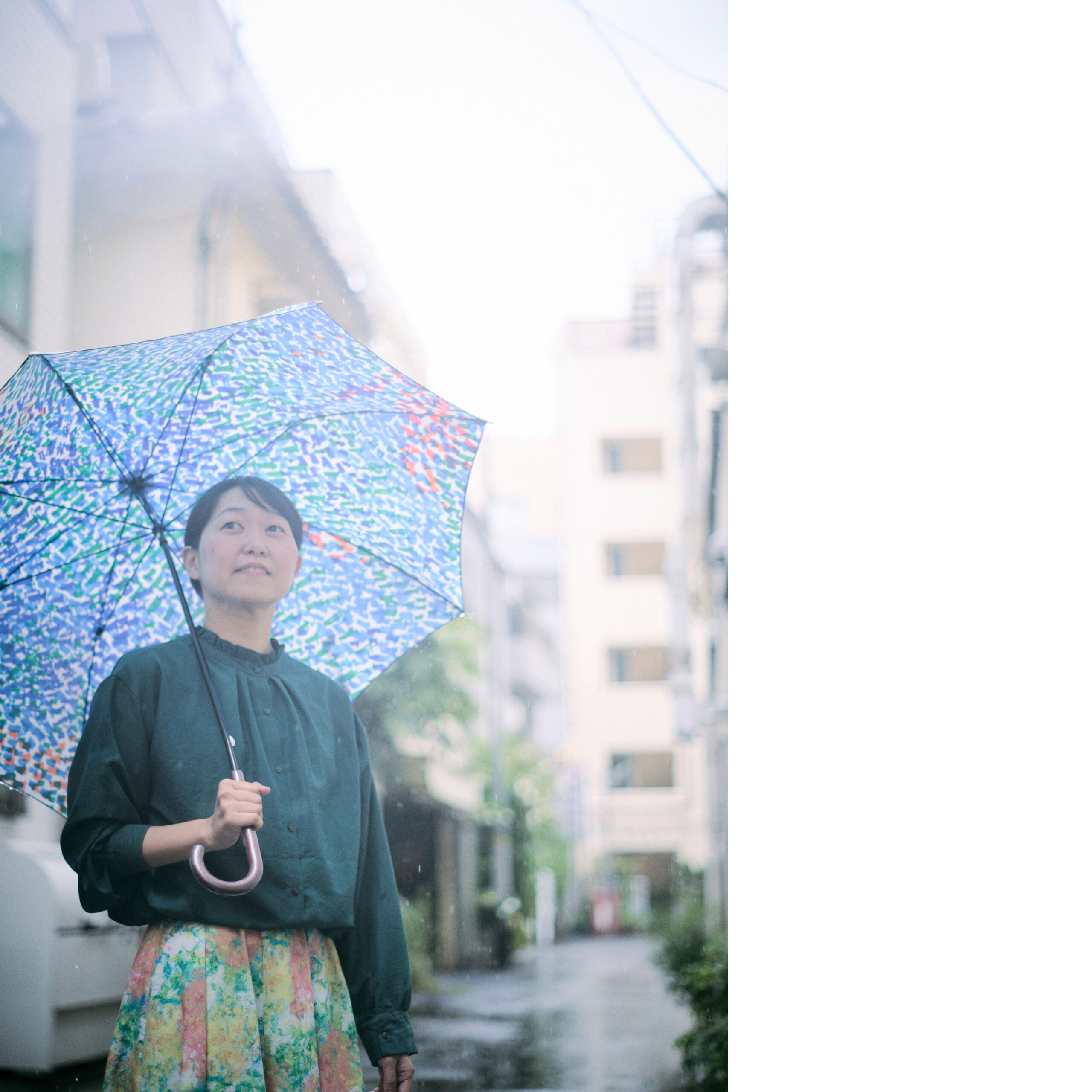 路地裏で傘をさして斜め上をみている菊永さんの写真
