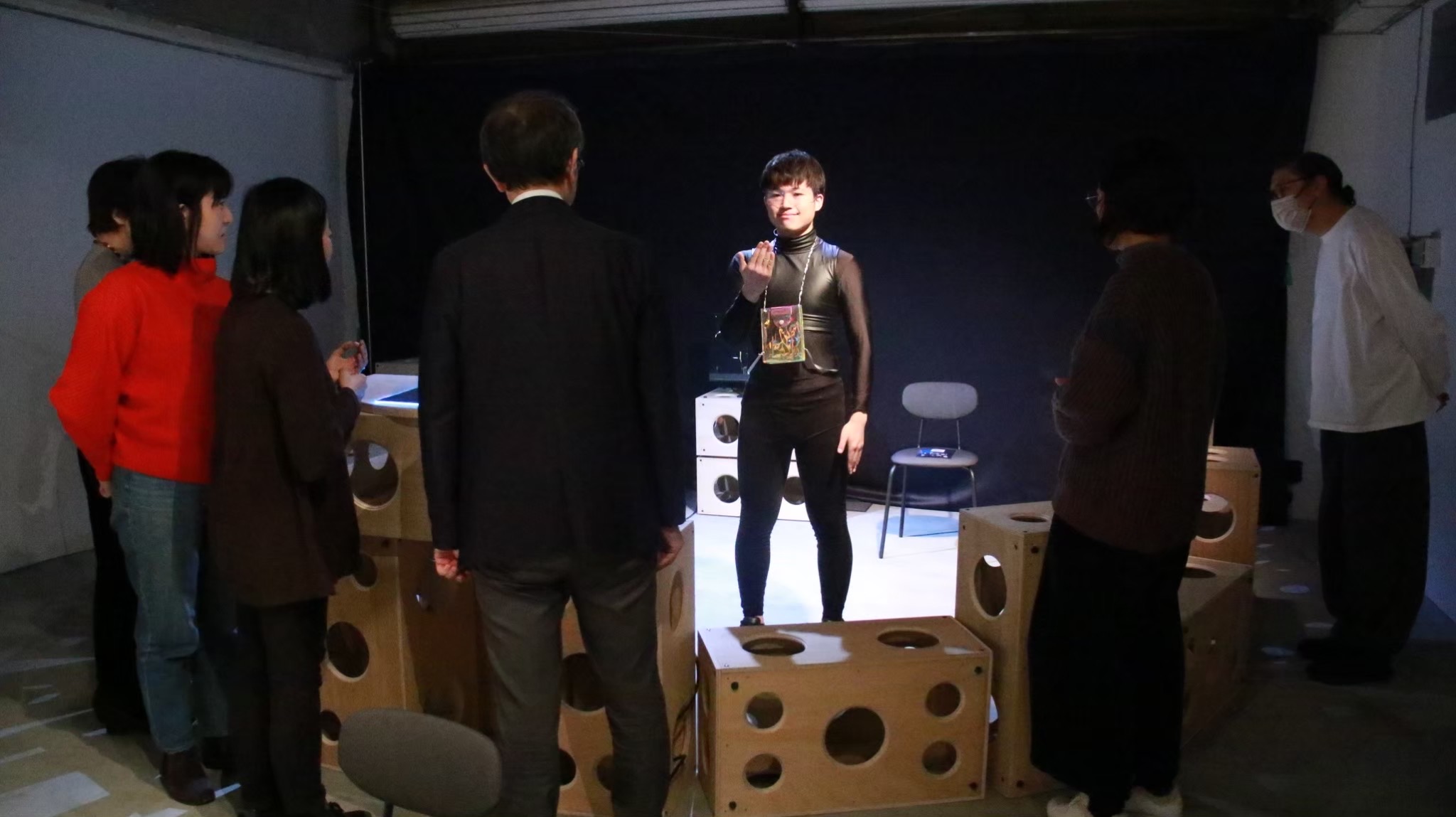 真ん中に黒い服を着て光を当てられた男性が立っており、周りに5名の人が男性をみている写真