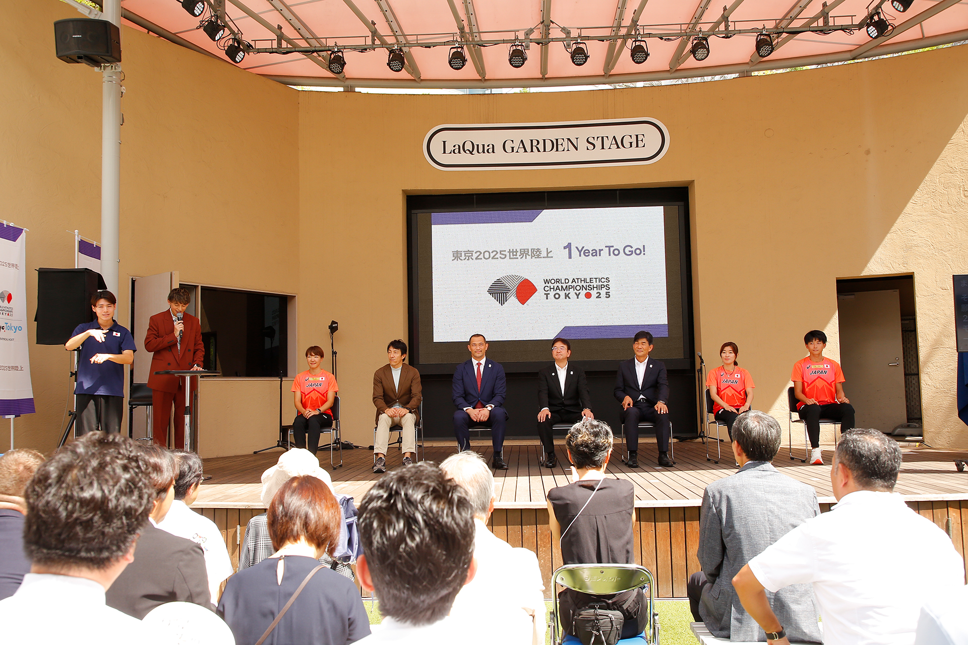 舞台上で登壇者7名が椅子に座っており、司会者のハリー杉山さんが話をしていて、その横で手話通訳がいる写真