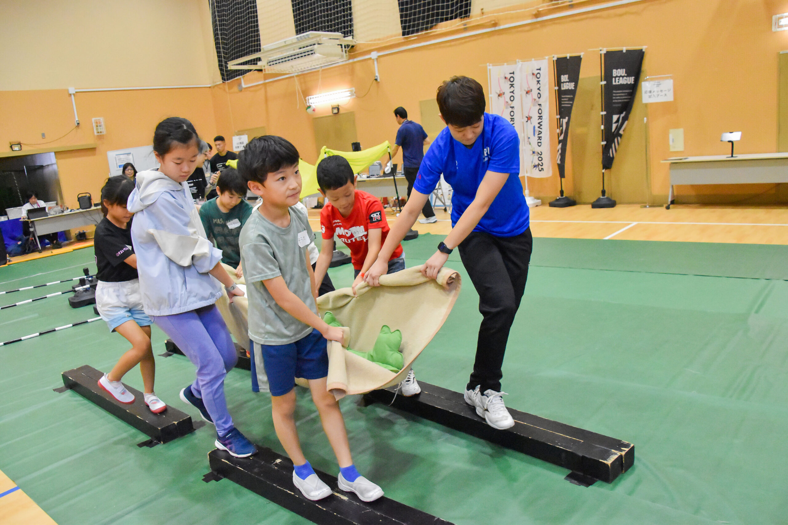 毛布にくるまれたぬいぐるみを細い木の上を歩き運んでいる中田選手と参加者の写真